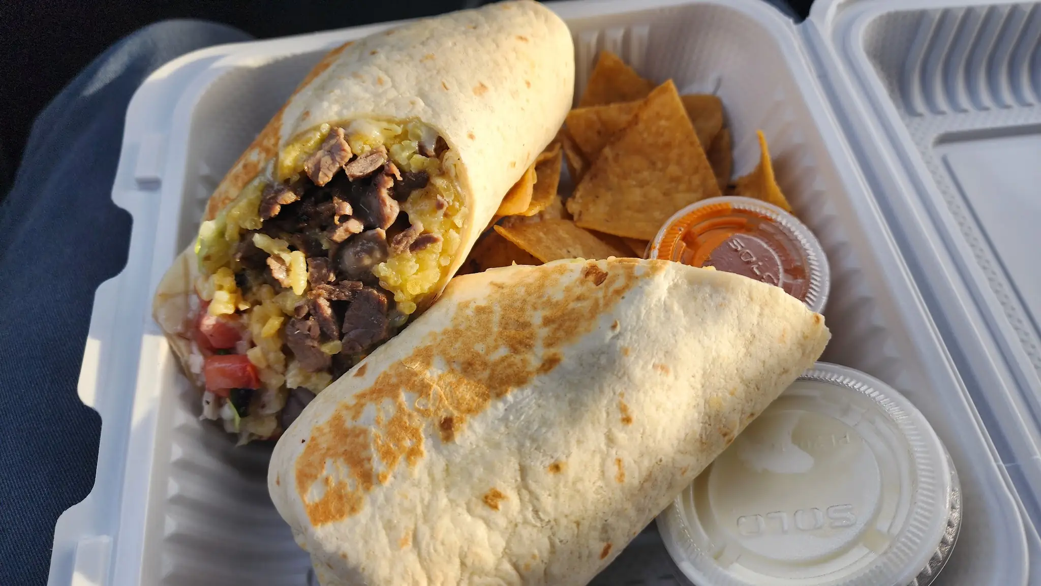 Burrito cut in half, filled with meat, rice and pico de gallo, with tortilla chips and salsa cups in a takeout box