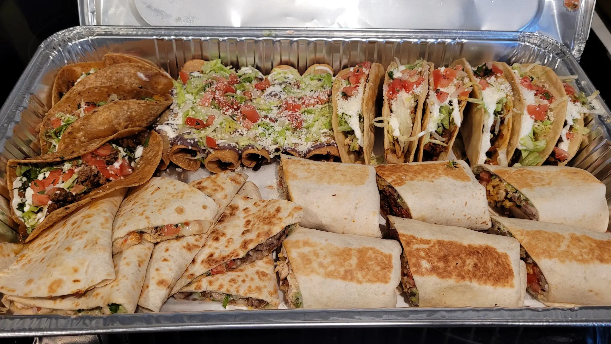 Foil tray of crispy tacos, tacos dorados topped with lettuce and crema, quesadilla wedges and burritos