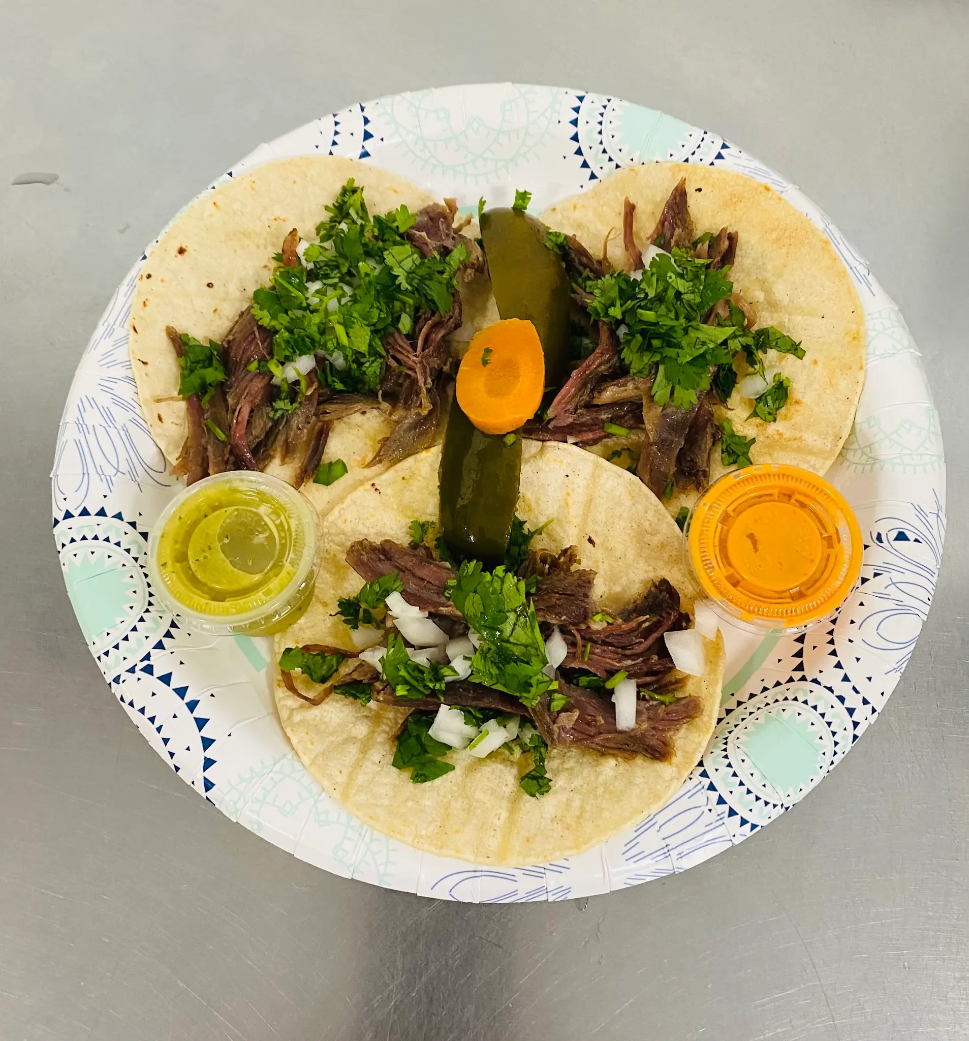 Three corn tacos with shredded meat, cilantro and onion, a pickled jalapeño and two cups of salsa on a paper plate