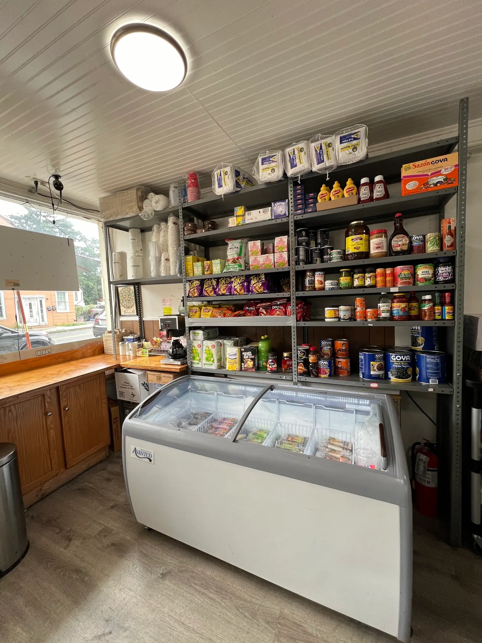 Steel shelves of Goya cans, hot sauces and Takis above a chest freezer, beside a wood counter and the front window